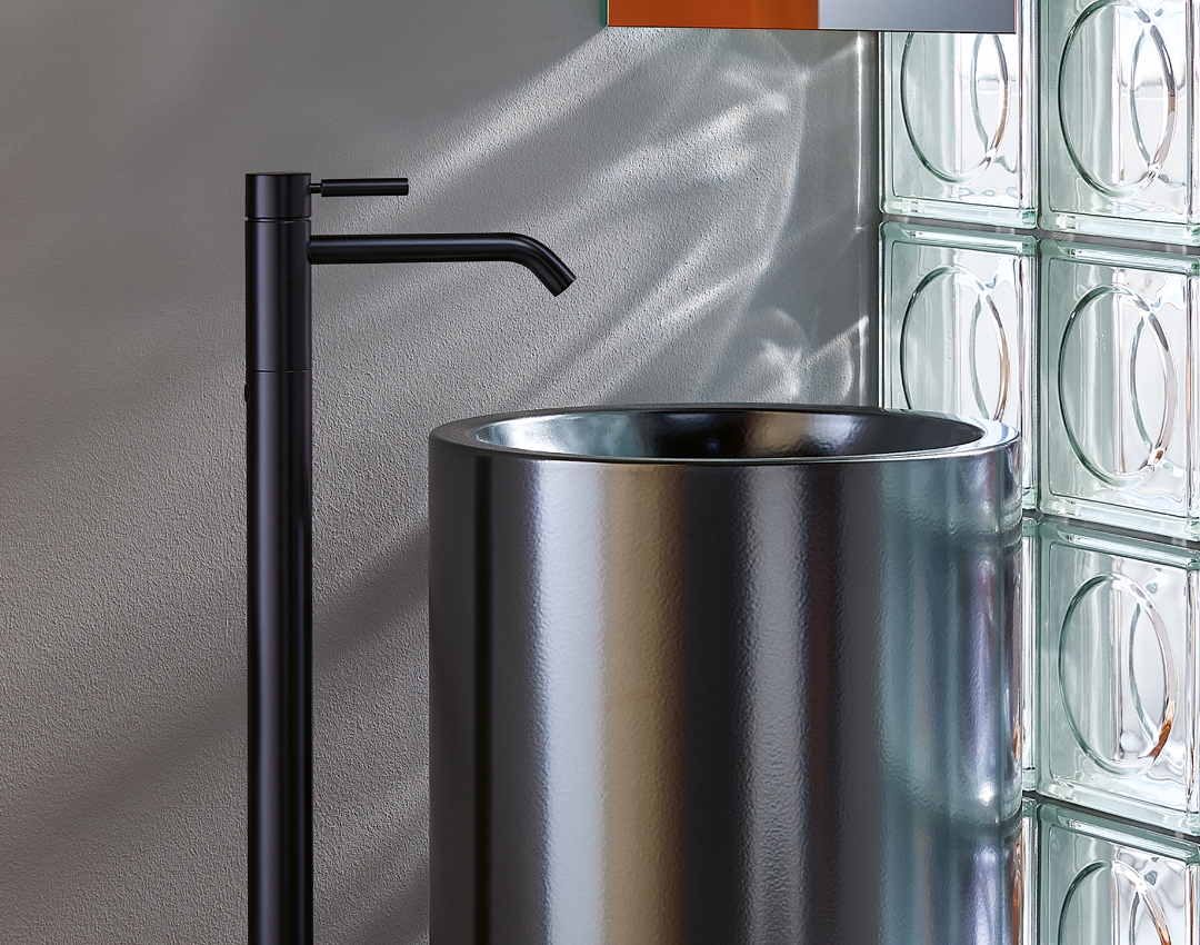 Dornbracht MEM floor-standing basin mixer in black matte finish, featured with a metallic cylindrical washbasin and glass block wall backdrop.